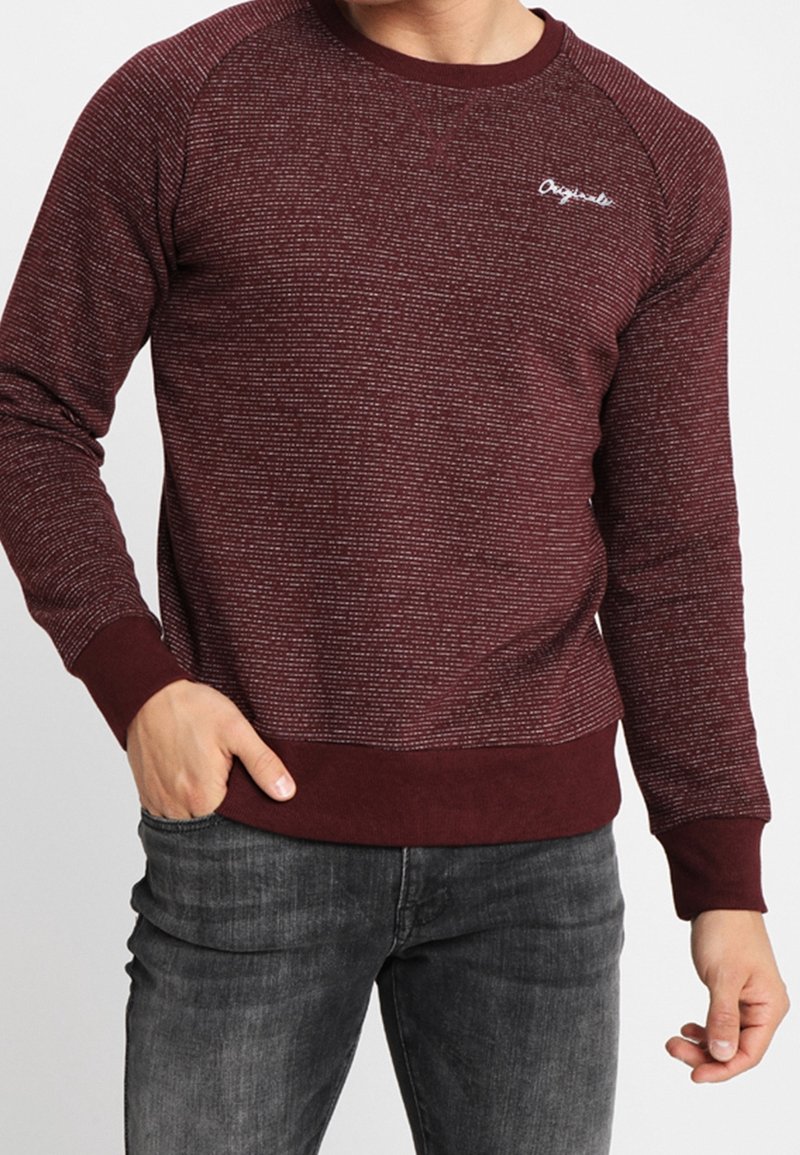 Homme portant un pull ajusté bordeaux texturé avec des poignets et un ourlet côtelés, associé à un jean gris foncé, main dans la poche.