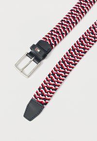 Spleciony pasek materiałowy w kolorze granatowym, czerwonym i białym z metalową klamrą w kolorze srebrnym oraz granatowymi skórzanymi wykończeniami, leżący płasko na białej powierzchni.