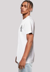 White cotton t-shirt with short sleeves, featuring a black graphic design on the left chest. Smooth texture, relaxed fit, worn with light denim jeans.
