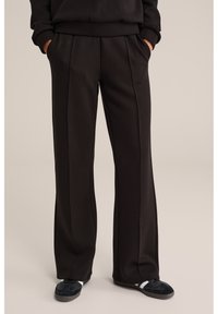 Persoană purtând pantaloni negri lejeri și adidași negri cu dungi albe, stând cu mâinile în buzunare pe un fundal neutru.