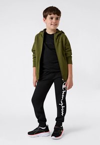 Casaco verde com fecho éclair, t-shirt preta, calças de treino pretas com logótipo branco da "Champion" e ténis pretos com detalhes vermelhos. Roupa casual atlética.