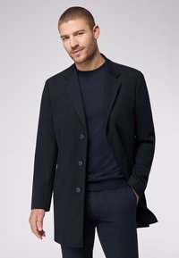 Cappotto nero su misura realizzato in tessuto liscio, con chiusura a un bottone, due bottoni, rever a punta e taschino sul petto. Indossato sopra un maglione scuro.