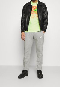 Černá kožená bomber bunda, jasně zelené tričko s grafikou, šedé jogger kalhoty s logem a černé tenisky. Jednoduchý, sportovní outfit.