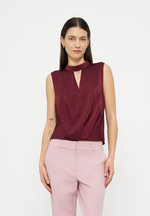 Γυναίκα με μακριά σκούρα μαλλιά φοράει αμάνικο μαρουνί μπλουζάκι με λαιμόκοψη τύπου «κλειδί» και ανοιχτό ροζ παντελόνια, στέκεται μπροστά σε απλό φόντο.