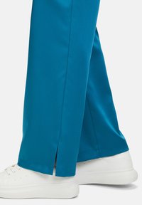 Turkosa satinbyxor med rak passform, med sidofickor vid anklarna, i kombination med vita sneakers. Slät yta och livfull färg.