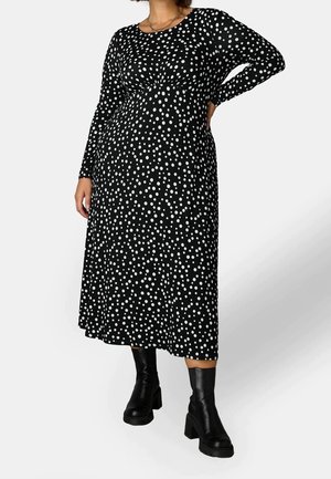 Kobieta w czarnej sukience midi w białe kropki z długimi rękawami, w połączeniu z czarnymi masywnymi botkami na obcasie.