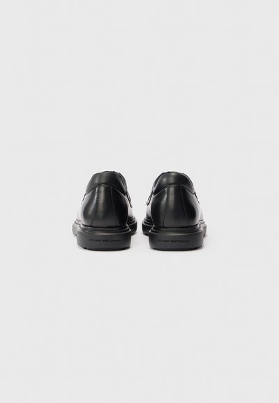Sorte læder dress sko set fra bagsiden, med "Stuart Weitzman" branding på sålen mod en simpel hvid baggrund.