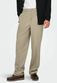 Beežid laiade säärtega püksid pidulike voltidega, vööaasad ja küljetaskud, mille juurde on kombineeritud pruunid paadijalanõud ja must kardigan. Materjaliks puuvillasegu.