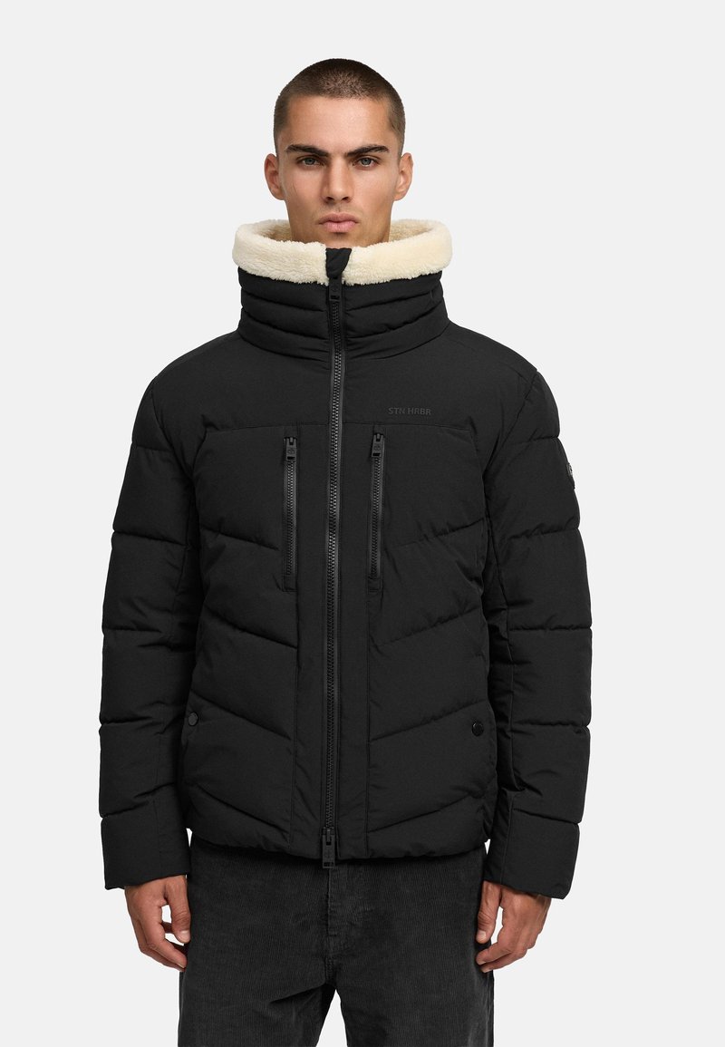 Melnā pūkainā jakā ar augstu apkakli, kas izklāta ar krēmkrāsas mākslīgo kažokādu, ar kabatām ar rāvējslēdzēju un quilted rakstu. Mīksta auduma struktūra.
