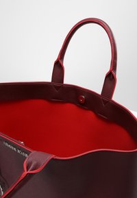 Czerwona torba tote z gładką, błyszczącą powierzchnią i kontrastowym czerwonym wnętrzem. Posiada solidne uchwyty oraz zapięcie na zatrzask, a w środku znajduje się detal z logo.