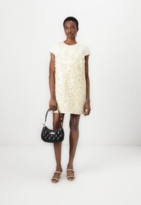 Short-sleeve, cream dress with floral embroidery, worn with a black quilted handbag and beige sandals, on a model against a plain background.