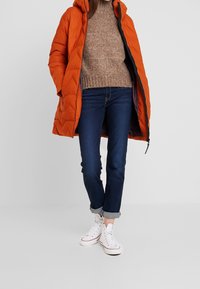 Manteau doudoune orange vif avec capuche et texture matelassée, pull en laine marron, jean bleu foncé, et baskets blanches avec des accents rouges.