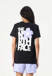 Črna bombažna majica s kratkimi rokavi, ki na hrbtni strani nosi veliko belo besedilo "THE NORTH FACE" in vijolično cvetlično grafiko. Ohlapen kroj.