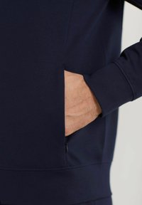 Suéter azul marino con una textura suave, presenta un bolsillo lateral y puños ajustados. El diseño enfatiza la comodidad y la practicidad.