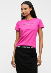 Tricou din bumbac roz aprins cu mâneci scurte, având textul "KARL LAGERFELD JEANS" în negru pe piept. Poartă o fustă neagră.