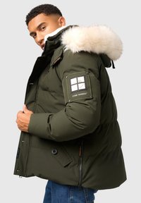 Zelená prešívaná bunda s kapucňou s kožušinovým okrajom, na rukáve s logom, predné vrecká a zapínanie na zips. Textúrovaný, prešitý dizajn.