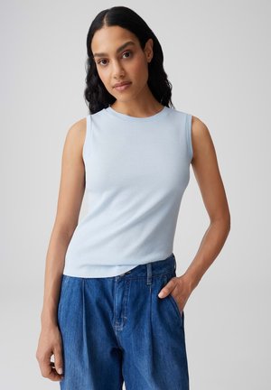 Femme aux longs cheveux noirs portant un débardeur bleu clair et un jean taille haute bleu, debout devant un fond gris uni.