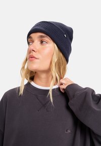 Donkerblauwe ribgebreide beanie met een omgeslagen rand. Draag het met een grijze trui met een klein logo. Eenvoudig, casual ontwerp met een gestructureerde afwerking.