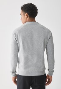 Man met krullend haar, gekleed in een lichtgrijze sweatshirt met lange mouwen en zwarte broek, staande tegen een effen witte achtergrond, met zijn rug naar de camera.