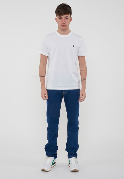 T-shirt bianca in cotone con maniche corte e un piccolo logo, abbinata a jeans denim blu e sneakers bianche con dettagli verdi.