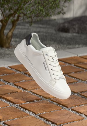 Baskets blanches en cuir avec un talon noir, design à lacets et branding "Calvin Klein" sur le côté, placées au-dessus de carrelage en bois avec du gravier.