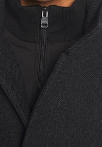 Gros plan sur un manteau noir texturé porté par-dessus une veste noire zippée avec un col côtelé et un tirette métallique gravé du mot « ESPRIT ».