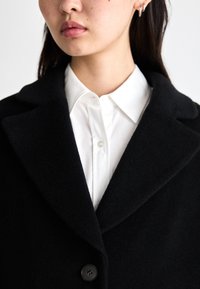 Schwarzer Wollmantel mit Reverskragen, ausgestattet mit einem einzigen Knopf und strukturiertem Stoff. Trägt man über einem weißen Hemd mit Kragen.