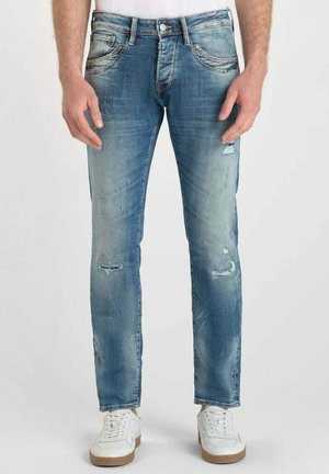 Jeans en denim bleu clair avec une coupe slim, présentant des détails déchirés et un effet décoloré. Le modèle porte des baskets blanches.