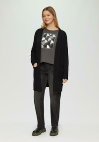 Zwarte cardi met lange mouwen, open voorkant en zakken; gedragen over een grijze graphic t-shirt met ronde patronen; gecombineerd met donkerblauwe jeans en chunky laarzen.