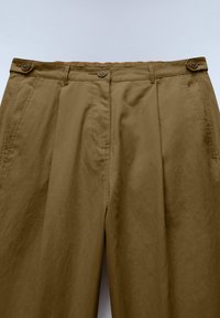 Braune High-Waist-Hose mit vorderen Falten, Seitentaschen, Gürtelschlaufen und Knopfdetails an Taille und Seiten.