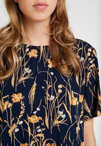 Marinblus med gult och vitt blommönster, med rund halsringning, korta ärmar och lätt, strukturerad tyg.