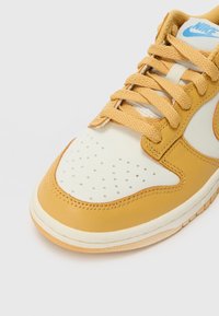 Nike-sneaker med en kremfarget lær overdel, gule lærdetaljer, perforert tå og runde lisser. Har en blå logo.