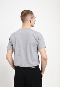 Šedé tričko s krátkým rukávem a hladkou texturou, v kombinaci s černými kalhotami. Tričko má kulatý výstřih a volný střih.