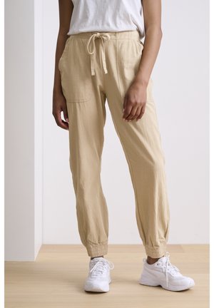 Personne portant un pantalon beige à cordon avec poches et poignets élastiques, associé à des baskets blanches sur un sol en bois devant un mur blanc.