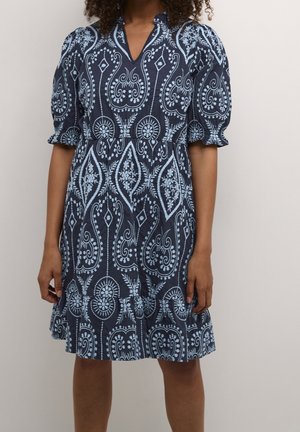 Femme portant une robe bleu marine aux genoux avec une broderie florale et géométrique complexe en bleu clair, des manches bouffantes et un col en V.