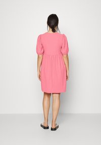 Donna con capelli scuri raccolti, che indossa un abito rosa al ginocchio con maniche a sbuffo, in piedi di fronte a un muro bianco semplice, con sandali con plateau neri.