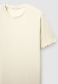 T-shirt de couleur crème avec un col rond, des manches courtes et une surface en tissu texturé. L'étiquette porte l'inscription "CALLIOPE".