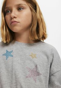 Szürke pamut póló, amelyen kék, sárga, rózsaszín és ezüst színű, strasszkőből készült csillag minták találhatók, valamint a "Designed" szó írásos formában.