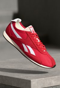 Crvene Reebok tenisice s bijelim prugama i vezicama, lebde iznad kamenite površine protiv zamućene tamne pozadine.