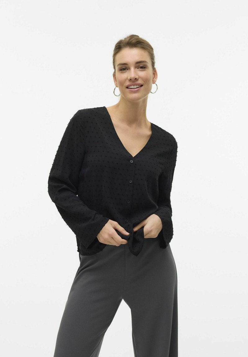Blusa nera con punti texture, scollo a V, maniche lunghe e chiusura con bottoni. Abbinata a pantaloni grigi a gamba larga per un look casual.