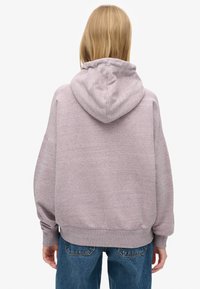 Oversize, kaptur z jasnoszarej tkaniny o fakturowanym wykończeniu. Posiada długie rękawy oraz ściągacz na dole, zestawiony z niebieskimi dżinsami.