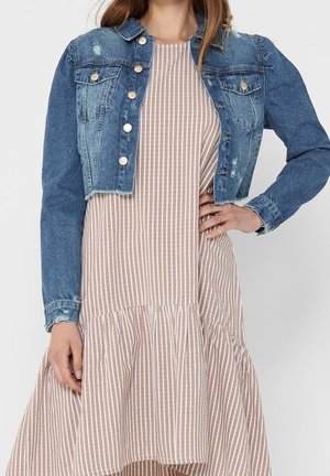 Vrouw draagt een cropped blauwe spijkerjasje over een beige-wit lang gestreepte jurk met ruches aan de zoom, hand op de heup.