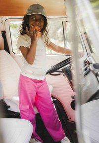 Witte geribbelde t-shirt met kleurrijke stippen, roze sweatpants met cargozakken, en een lichtblauwe pet, staand in een voertuiginterieur.