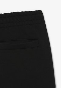 Zwarte trainingsbroek met een gladde textuur, elastische tailleband en een klein achterzakje. Heeft eenvoudige stiksels langs de tailleband.