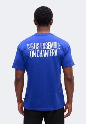 Homme portant un T-shirt bleu avec le texte blanc "TOUS ENSEMBLE ON CHANTERA" et un logo stylisé, vu de dos.
