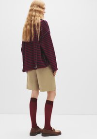 Gestreiftes, übergroßes Oberteil in Marineblau und Rot, beigefarbene Shorts und kniehohe kastanienfarbene Socken. Braune Schuhe mit niedrigem Profil und strukturiertem Design.