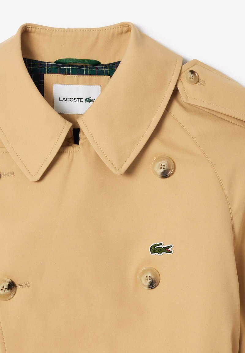 Col de trench beige avec boutons croisés, épaulettes, et logo crocodile vert de Lacoste sur la poitrine.