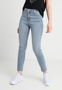 Persona vestida con unos pantalones vaqueros ajustados de tiro alto de color azul claro y zapatillas blancas, de pie contra un fondo claro y liso.