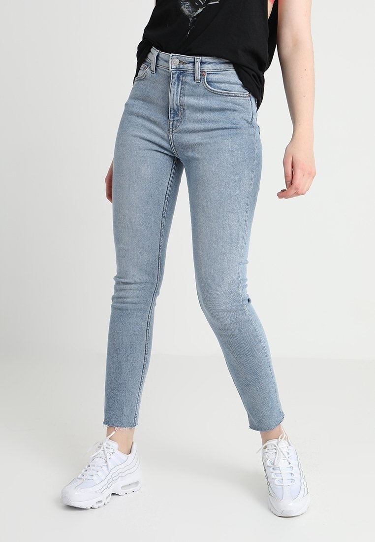 Persona vestida con unos pantalones vaqueros ajustados de tiro alto de color azul claro y zapatillas blancas, de pie contra un fondo claro y liso.