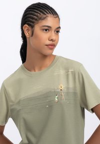 Olive green cotton t-shirt featuring a graphic illustration of a windmill and a person, with a relaxed fit and short sleeves.